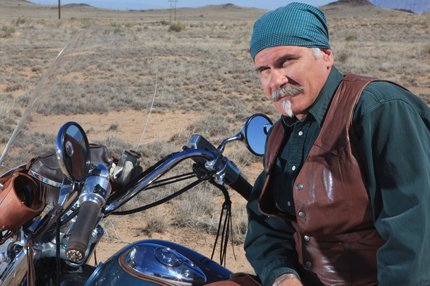 knappe oudere man leunend tegen grote motorfiets buitenshuis in bruin woestijn - Foto, afbeelding