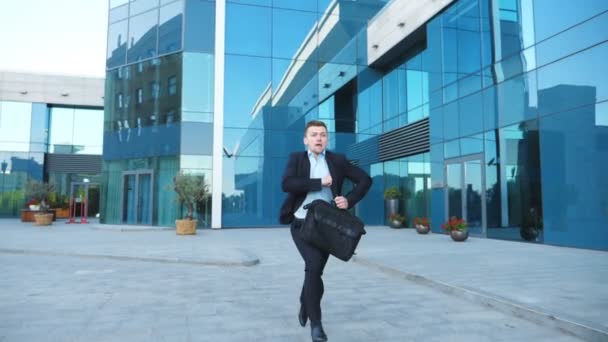 Young businessman with briefcase runs and looks at his watch. Confident guy late for meeting. Successful man in suit jogging near modern building. Handsome man in hurry to appointment. Slow motion - Footage, Video
