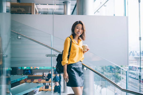 Happy Asian international student with leather backpack rejoicing indoors holding coffee takeaway cup and smiling at camera while waiting university friend, concept of happiness and joyful mood - Photo, image