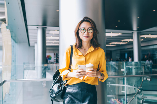 Half length portrait of attractive female blogger in optical spectacles for provide eyes protection smiling at camera during coffee break for networking with cellphone gadget, millennial person - Foto, afbeelding