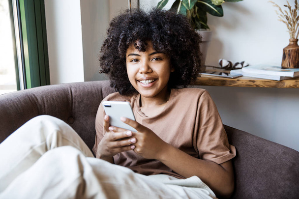 Beautiful smiling african american woman using smartphone in armchair at home - Photo, Image
