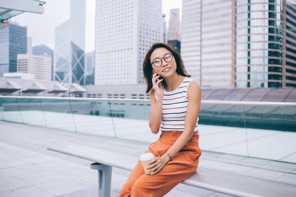 Positive female tourist with caffeine beverage taking rest at urban setting receiving smartphone cell call from friend for talking and communicating, concept of modern technology and generation z - 写真・画像