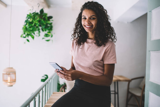 Half length portrait of dark skinned young woman looking at camera while sitting with cellphone indoors, African American smiling hipster girl holding modern mobile phone in hands during rest - Foto, Imagem