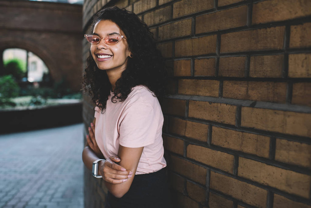Half length portrait of cheerful female student dressed in gorgeous pink sunglasses posing near brick wall with crossed hands, happy African American woman 20s enjoying good day on city street - Foto, afbeelding