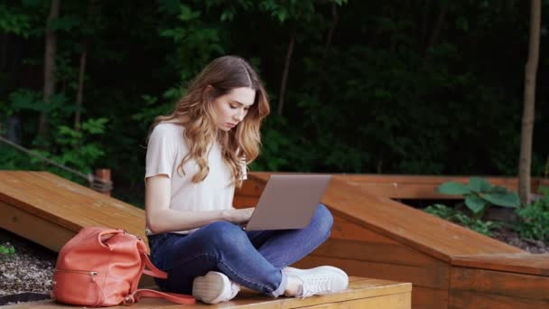 Smiling brunette woman in casual clothes using laptop computer while sitting on bench outdoors - Metraje, vídeo