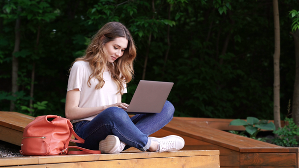 Happy brunette woman in casual clothes using laptop computer while sitting on bench outdoors - Imágenes, Vídeo