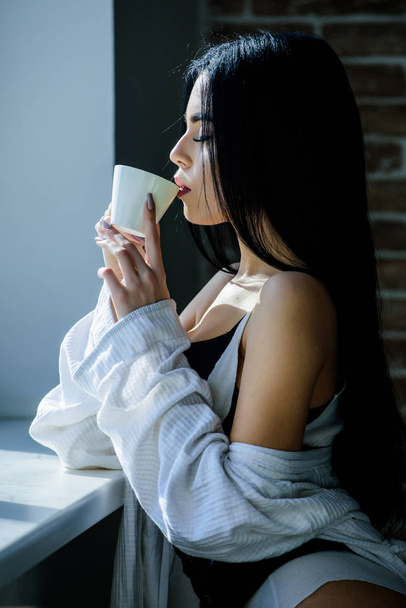 The best coffee for her daily routine. Sensual girl drinking her favorite morning coffee. Pretty woman drinking fresh hot coffee at window. Sexy girl holding ceramic coffee cup - Фото, изображение