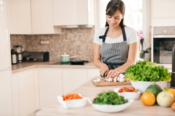 Beautiful smiling dedicated Caucasian brunette in apron standing in kitchen and chopping mushrooms. On table are lots of vegetables. Cooking at home concept. - Foto, Imagem