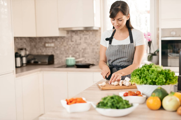 Beautiful smiling dedicated Caucasian brunette in apron standing in kitchen and chopping mushrooms. On table are lots of vegetables. Cooking at home concept. - Photo, image