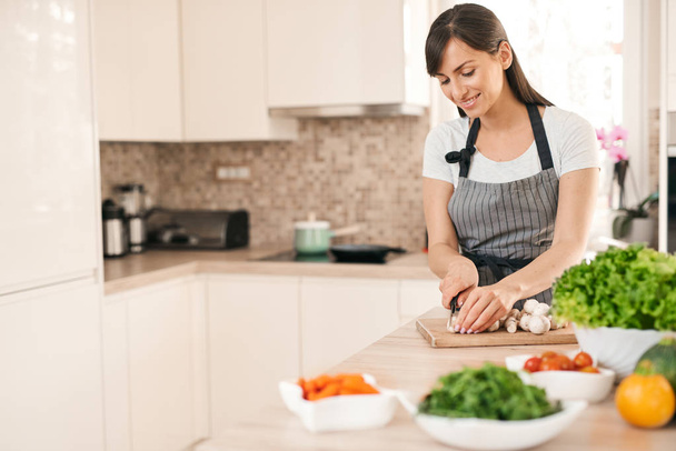 Beautiful smiling dedicated Caucasian brunette in apron standing in kitchen and chopping mushrooms. On table are lots of vegetables. Cooking at home concept. - Fotoğraf, Görsel