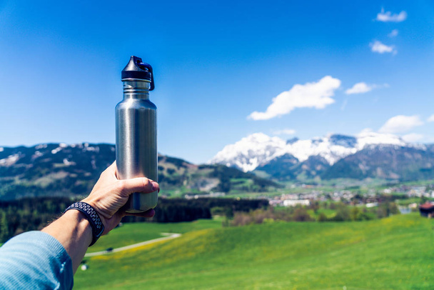 Male hand holding metal water bottle. Mountains and greenery in  - Фото, изображение