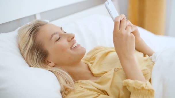 Beautiful blonde woman wearing pajama using phone in her bedroom.  - Πλάνα, βίντεο