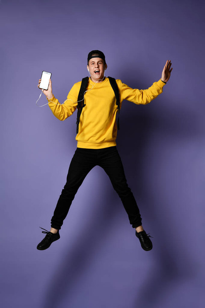 Handsome young student man in casual wear with backpack and headphones is listening to music using a smartphone and smiling while jumping - Foto, Imagen