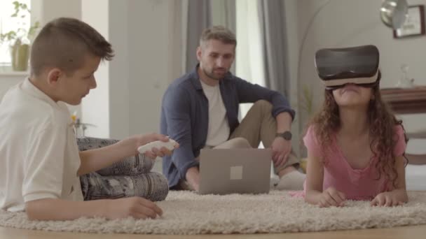 Brother and sister laying on the floor on the fluffy carpet using virtual reality glasses, father sitting on the background with children. Happy friendly family at home - Materiał filmowy, wideo
