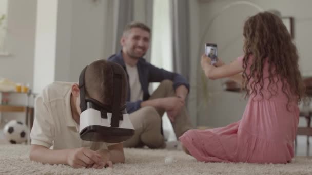 The boy and girl laying on the floor on fluffy carpet, the brother using virtual reality glasses, sister taking photo of father who sitting on the background of children. Happy friendly family at home - Video
