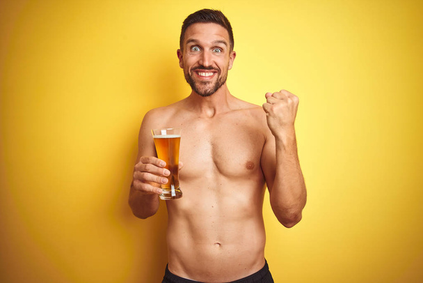 Jovem bonito homem sem camisa bebendo uma caneca de cerveja sobre fundo amarelo isolado gritando orgulhoso e celebrando vitória e sucesso muito animado, animando emoção
 - Foto, Imagem