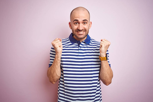 Jeune homme chauve à la barbe portant un t-shirt bleu rayé décontracté sur fond rose isolé célébrant surpris et étonnés du succès avec les bras levés et les yeux ouverts. Concept gagnant
. - Photo, image