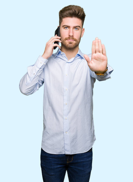 Молодий красивий чоловік розмовляє на смартфоні з відкритою рукою робить знак зупинки з серйозним і впевненим виразом, жест оборони
 - Фото, зображення