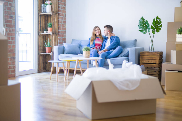 junges schönes Paar, das auf dem Sofa in der neuen Wohnung sitzt und sich umarmt, mit Pappkartons um sie herum, sehr aufgeregt für den Umzug in ein neues Zuhause - Foto, Bild