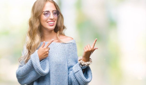 Beautiful young blonde woman wearing sunglasses over isolated background smiling and looking at the camera pointing with two hands and fingers to the side. - Photo, Image