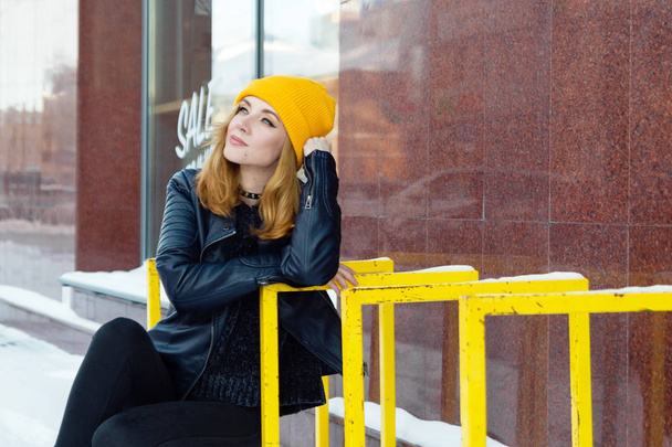 Young woman with blue eyes and blond hair in a yellow knitting hat and black leather jacket on the snowy street of city. - Photo, Image