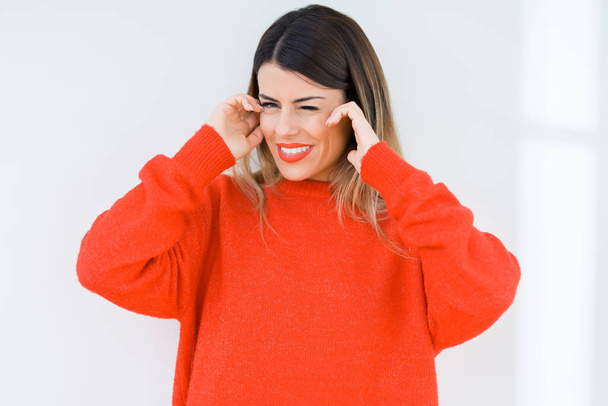Młoda kobieta ubrana casual czerwony sweter na białym tle obejmujące uszy z palcami z irytują wyraz hałasu głośnej muzyki. Głuchy koncepcja. - Zdjęcie, obraz