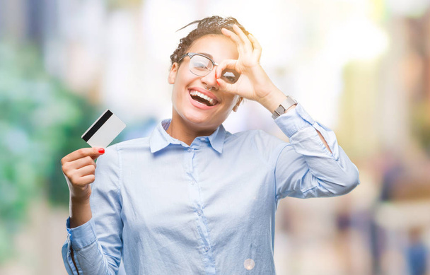 junge afrikanisch-amerikanische Mädchen hält Kreditkarte über isolierten Hintergrund mit glücklichem Gesicht lächelnd tut ok Zeichen mit Hand auf Auge durch Finger schauen - Foto, Bild