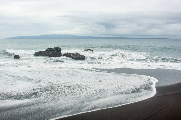 白い泡の波が岩と黒い砂浜に衝突, 遠くにヌサペニダとパンタイワテスで.バリ島 - 写真・画像