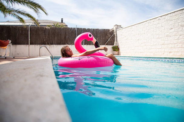 Jovem flutuando em um flamingo inflável em uma piscina azul um dia ensolarado de férias de verão
 - Foto, Imagem