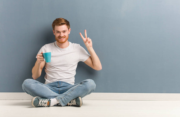 Jeune étudiant rousse assis sur le sol amusant et heureux de faire un geste de victoire. Il tient une tasse de café.
. - Photo, image