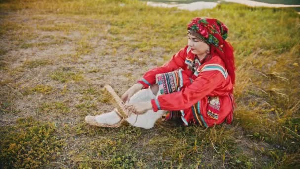 Nainen venäläisissä kansanvaatteissa laittaa bast kengät - istuu kentällä
 - Materiaali, video