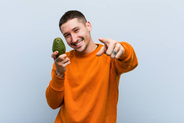 Young caucasian man holding an avocado cheerful smiles pointing to front. - Foto, immagini
