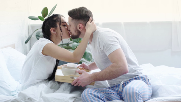 寝室でテレワークしながらラップトップを使用して陽気な女性、彼氏は朝食とキスのトレイを保持し、ガールフレンドにキス  - 映像、動画