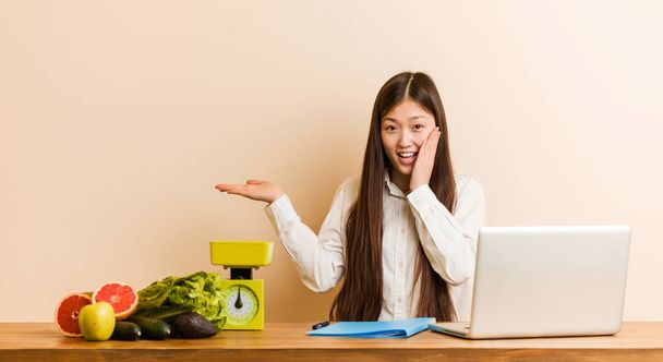 Young nutritionist chinese woman working with her laptop holds copy space on a palm, keep hand over cheek. Amazed and delighted. - Photo, Image