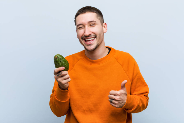 Jovem caucasiano segurando um abacate sorrindo e levantando o polegar
 - Foto, Imagem