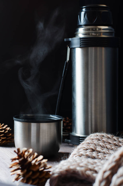 Metal thermos, hot tea or coffee in a mug. Brown knitted scarf. Autumn mood. Brown wooden background. Copy space - Foto, Bild