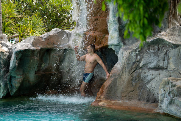 A young guy stands under artificial waterfalls in the pool on site - Photo, Image