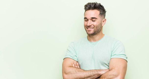 Jonge knappe man tegen een groene achtergrond lachend vertrouwen met gekruiste armen. - Foto, afbeelding
