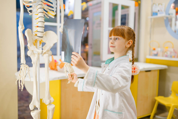 Маленькая девочка в форме смотрит на рентген, играет в доктора, детскую. Ребенок играет медицинского работника в воображаемой больнице, профессиональное обучение, детская мечта
 - Фото, изображение