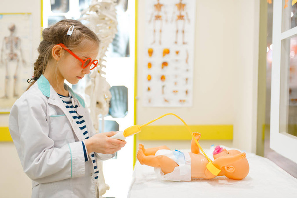 Маленькая девочка в форме играет в доктора с игрушечной куклой, игровая комната. Ребенок играет медицинского работника в воображаемой больнице, профессиональное обучение, детская мечта
 - Фото, изображение