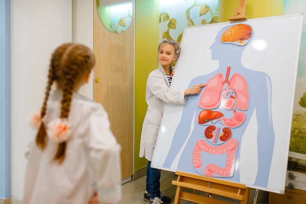 Bambine in uniforme che giocano al dottore al poster con organi umani, sala giochi. Bambini gioca medico operaio in ospedale immaginario, professione di apprendimento, sogno infantile
 - Foto, immagini