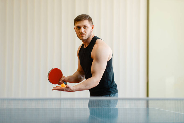 Man met pingpong racket voorbereidingen om een bal te raken, trainen binnenshuis. Mannelijke persoon in sportkleding, training in tafeltennis club - Foto, afbeelding