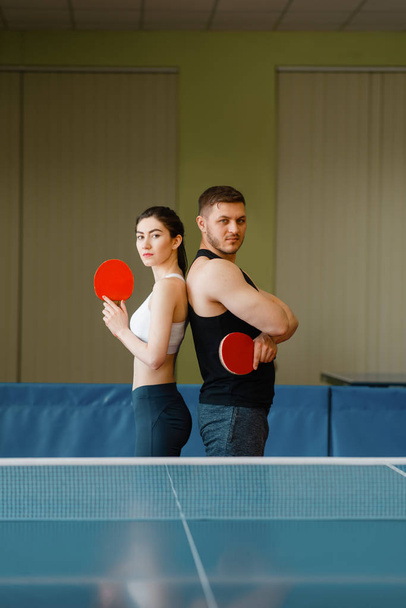 Paar mit Schlägern posiert an der Tischtennisplatte mit Netz drinnen. Mann und Frau in Sportbekleidung, Tischtennis-Trainingsspiel im Fitnessstudio - Foto, Bild