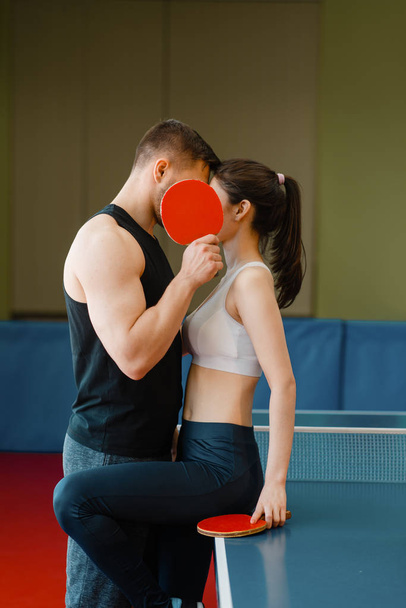 Casal com raquetes beijando na mesa de pingue-pongue com rede dentro de casa. Homem e mulher em sportswear, jogo de treinamento de tênis de mesa no ginásio
 - Foto, Imagem