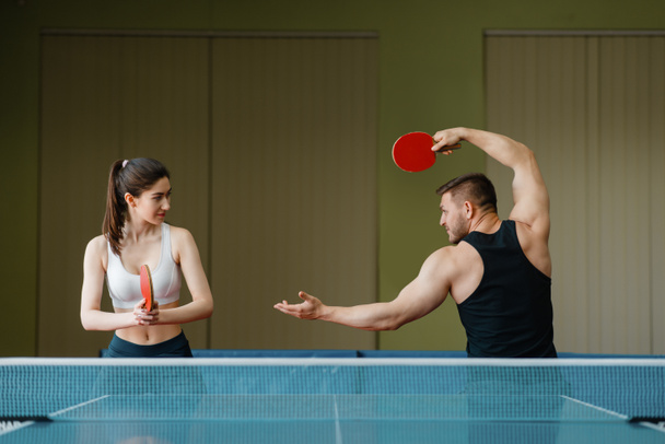 Hombre y mujer en entrenamiento de ping pong en interiores. Pareja en ropa deportiva sostiene raquetas y juega al tenis de mesa en el gimnasio. Hombres y mujeres en el club de tenis de mesa
 - Foto, Imagen