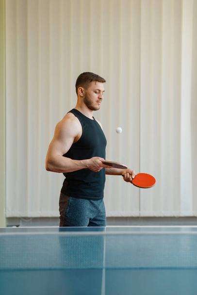 Człowiek z dwóch rakiet spożywczych ciosy w ping ponga w pomieszczeniu. Męska osoba w odzieży sportowej przy stole z siatką, trening w klubie tenis stołowy - Zdjęcie, obraz