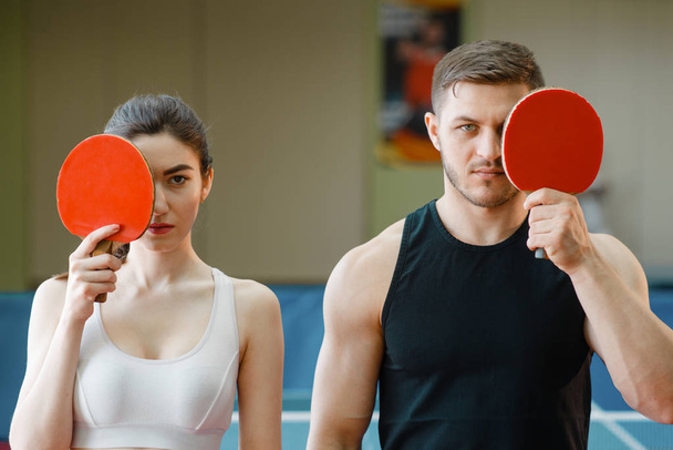 Uomo e donna tengono racchette da ping pong in casa. Coppia in abbigliamento sportivo gioca a ping pong in palestra. Persone di sesso maschile e femminile nel club di tennis da tavolo - Foto, immagini
