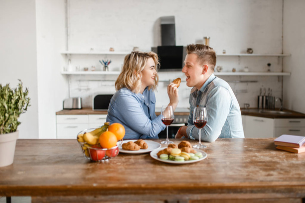 Attractive love couple sitting at the table, romantic dinner. Man and woman drinks wine and eats fruits on the kitchen. Happy lifestyle, beautiful relationship - Fotó, kép