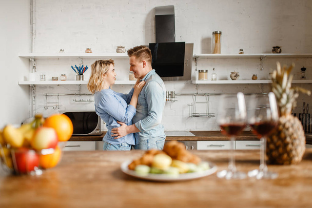 Two glasses of red wine on the table, love couple hugging on background, romantic dinner concept. Man and woman on the kitchen together. Happy lifestyle, beautiful relationship - Фото, изображение
