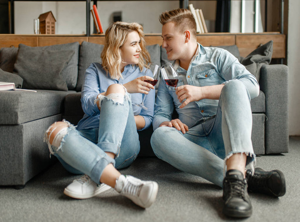 Young love couple sitting on the floor at the couch and drinks red wine, romantic dinner. Man and woman relaxing in living room. Happy lifestyle, beautiful relationship - Фото, изображение